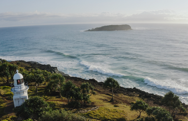 Lighthouse stands on a green mountain, with golden beaches, turquoise waters, and lush landscapes under a blue sky - The Tweed Coast