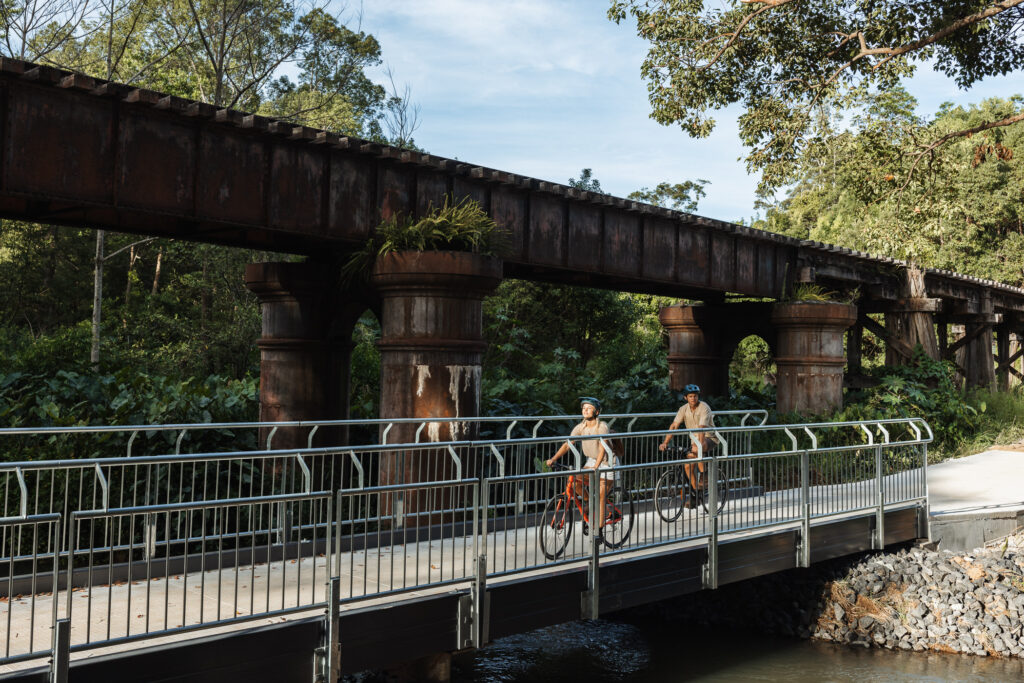 Couple riding bikes on Northern Rivers Rail Trail passed old railway bridge