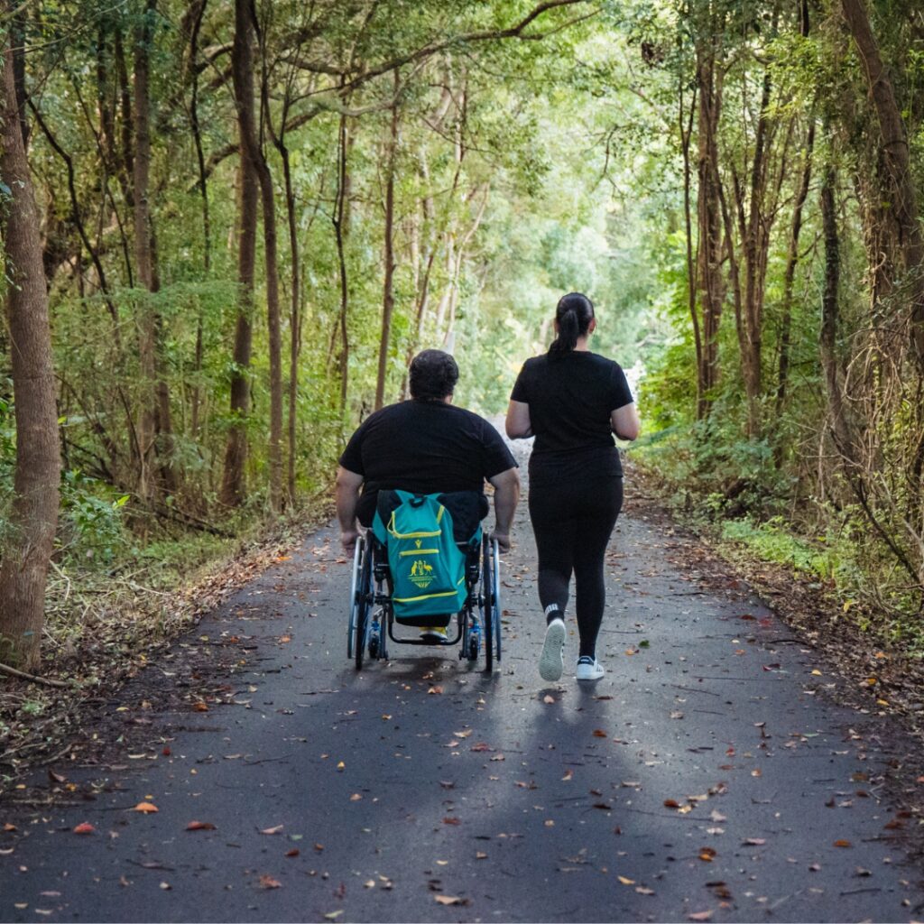 A couple on the Northern Rivers Rail Trail surrounded by lush trees: the man is in a wheelchair, and the woman walks beside him. They are enjoying the scenic, tree-lined path together.