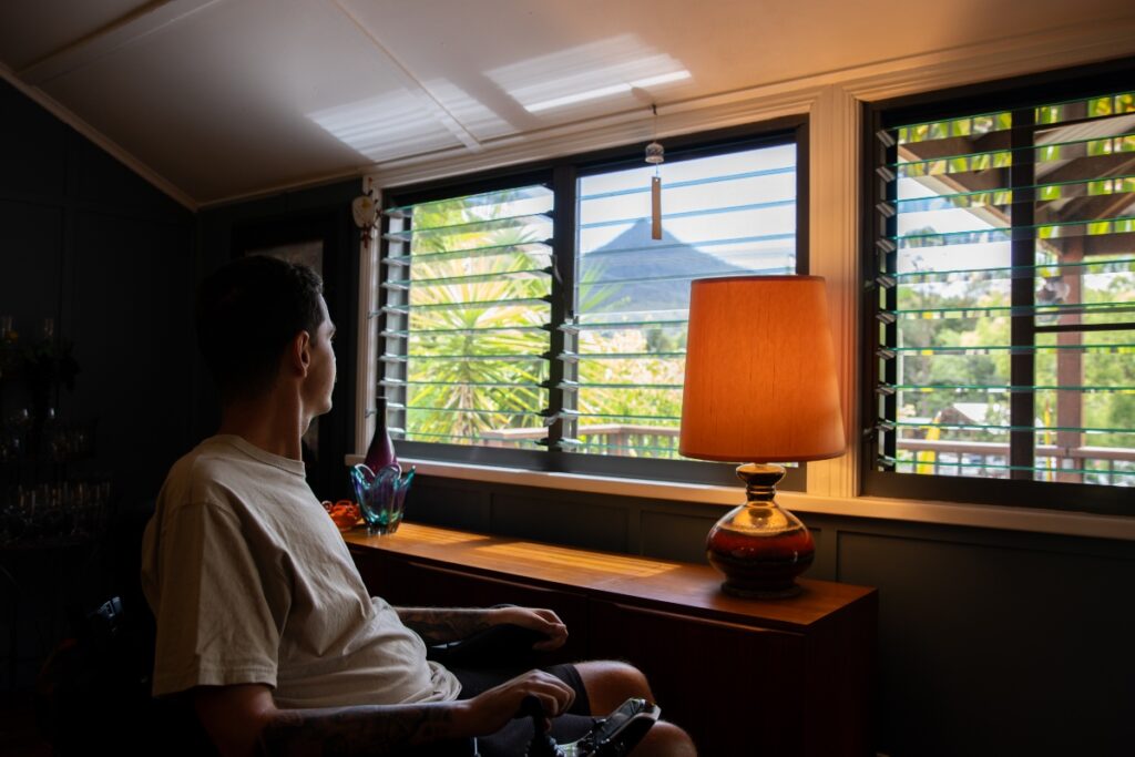 A man in a wheelchair gazes out of an accommodation window at the scenic view of Wollumbin (Mount Warning), with lush greenery and the mountain's silhouette in the distance.