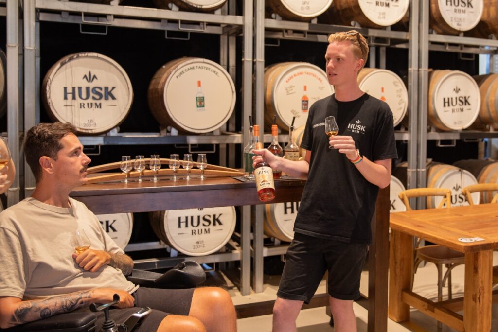 A man in a wheelchair enjoys a rum tasting during a distillery tour: he is holding a tasting glass and interacting with a guide, with distillery equipment visible in the background.