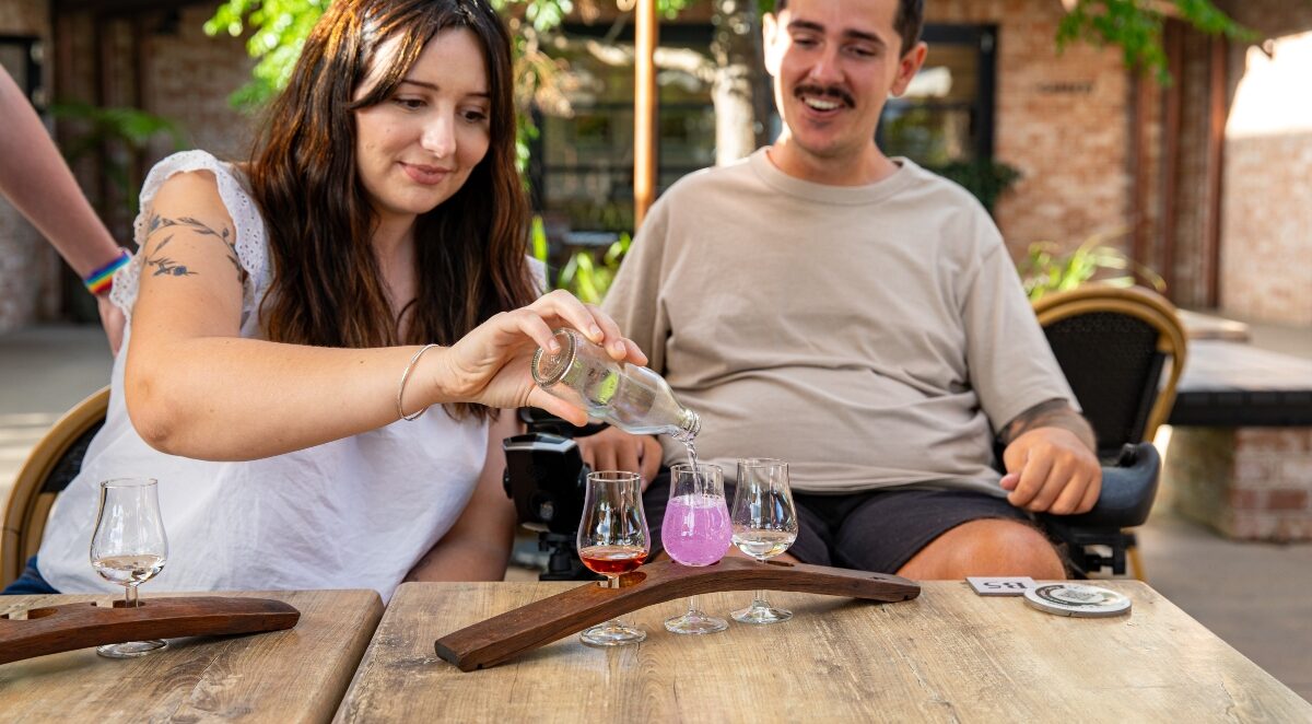 Accessible travel guide banner: A smiling couple at a distillery: the lady, seated, pours gin from a bottle. The man, also smiling, is seated in a wheelchair beside her, enjoying the moment.